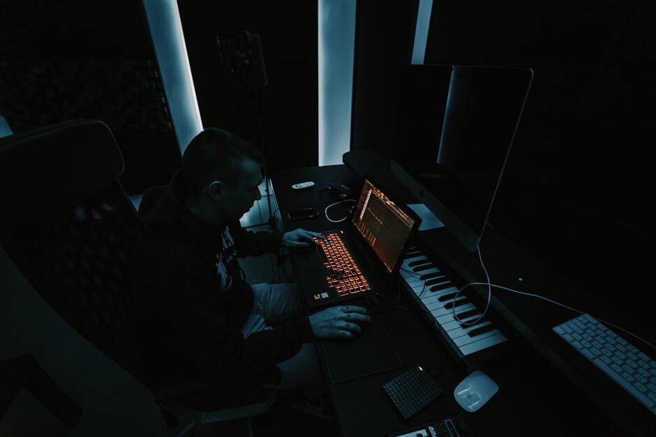 A music producer working at a mixing setup in a dark studio, orange keyboard glow and MIDI keyboard visible on a dimly lit desk