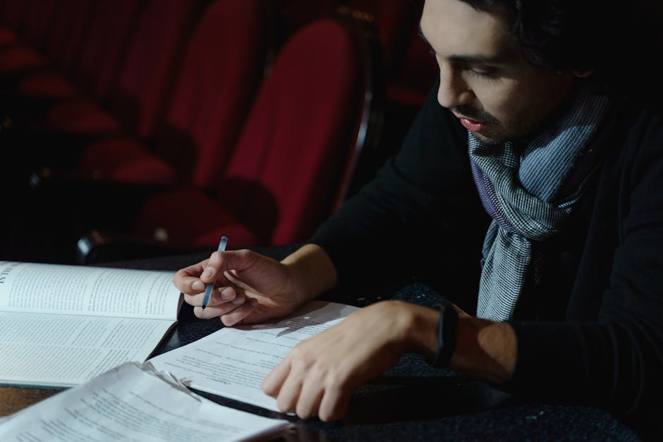 A writer reviewing multiple script drafts at a desk in a dimly lit theater auditorium