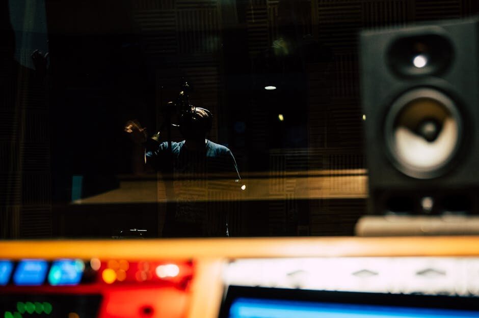 Silhouette of a vocalist recording behind glass in a dimly lit studio with monitor speakers