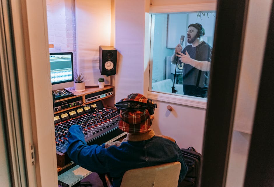 Audio engineer at a mixing console in a recording studio, with an actor visible through the glass booth window