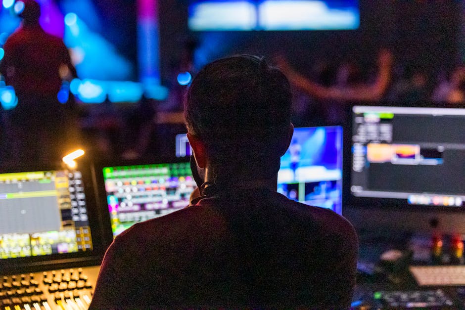 Production professional silhouetted against multiple glowing monitors in a darkened control room