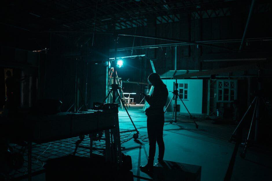 Grip crew member working on a large dark film sound stage with production lighting rigs visible in background