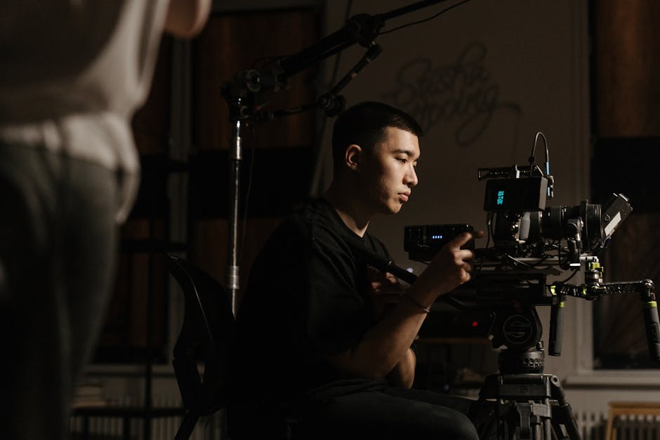 Camera operator seated at a film camera adjusting controls during a production, dark moody lighting