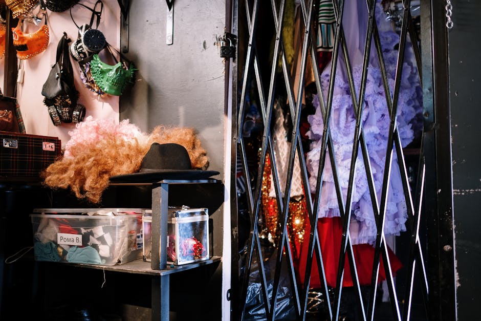 A theatrical costume department storage room with colorful period costumes, hats, and wigs hanging on racks behind a metal gate