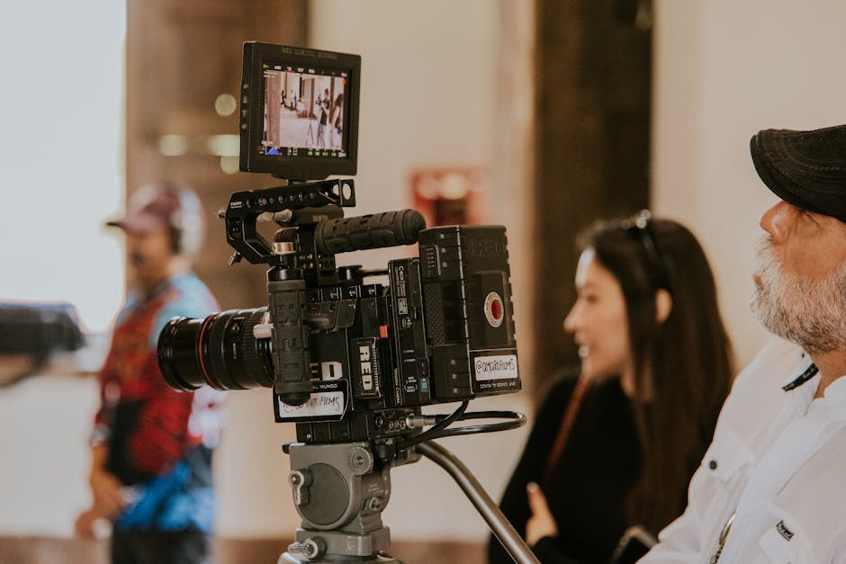 A RED cinema camera on a tripod with a cinematographer and crew visible in the background on a film set
