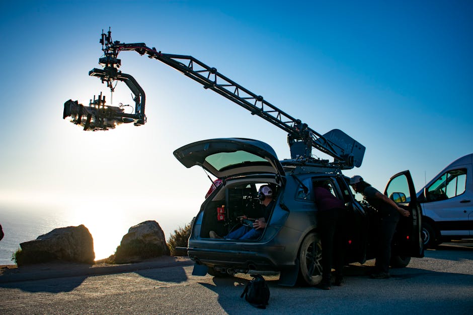 Film crew operating a camera crane mounted on a vehicle at a cliffside location during golden hour