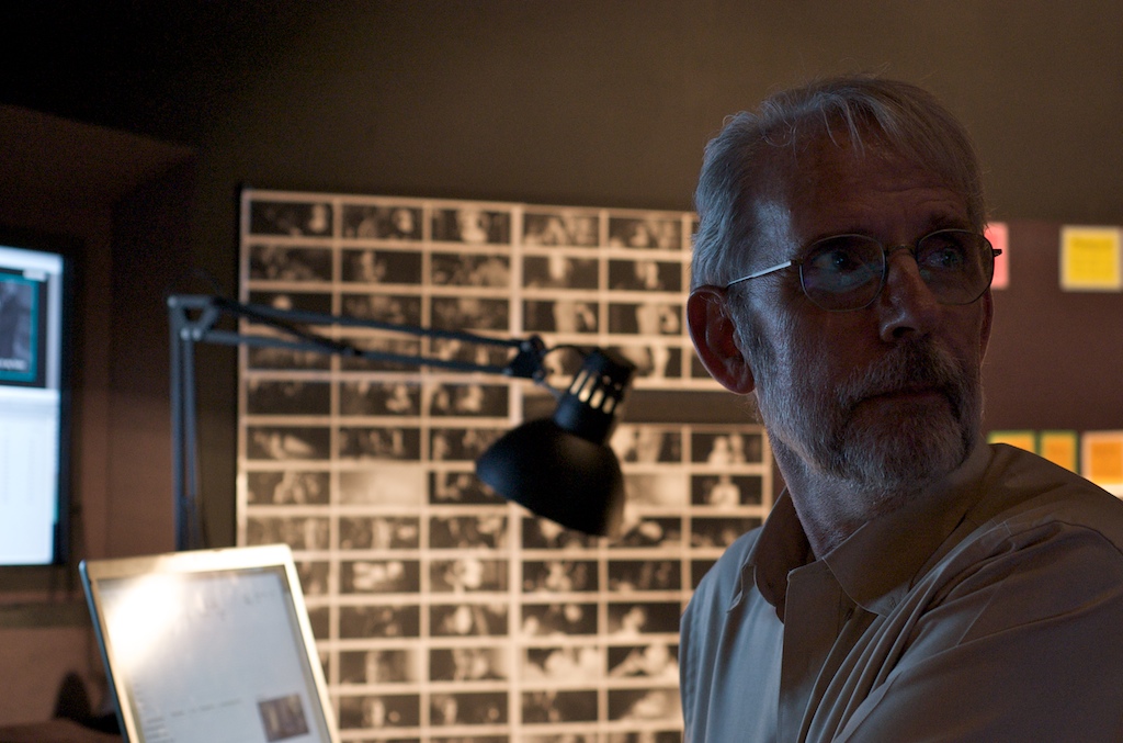 Walter Murch working at an editing station in a dark studio, film frame contact sheets pinned to wall behind him