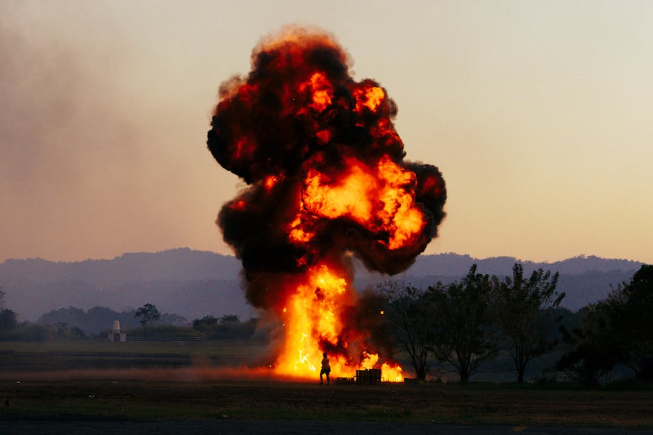 Large controlled pyrotechnic explosion with fire and smoke in an open field at sunset with a silhouetted figure nearby
